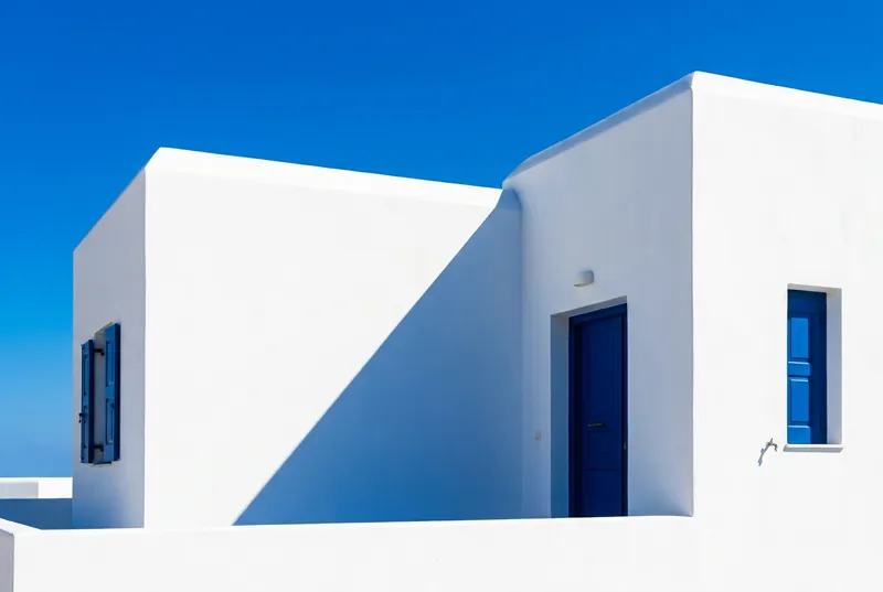A white cubic house with a deep cobalt-blue door and blue shutter, hard diagonal shadow.