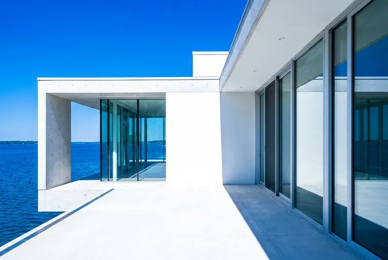 A white modern lakeside house with floor-to-ceiling glass beside deep blue water.